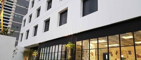 Ein modernes Gebäude mit weißer Fassade, zahlreichen Fenstern und einem Glasbereich am Eingang mit Stühlen und Tischen.