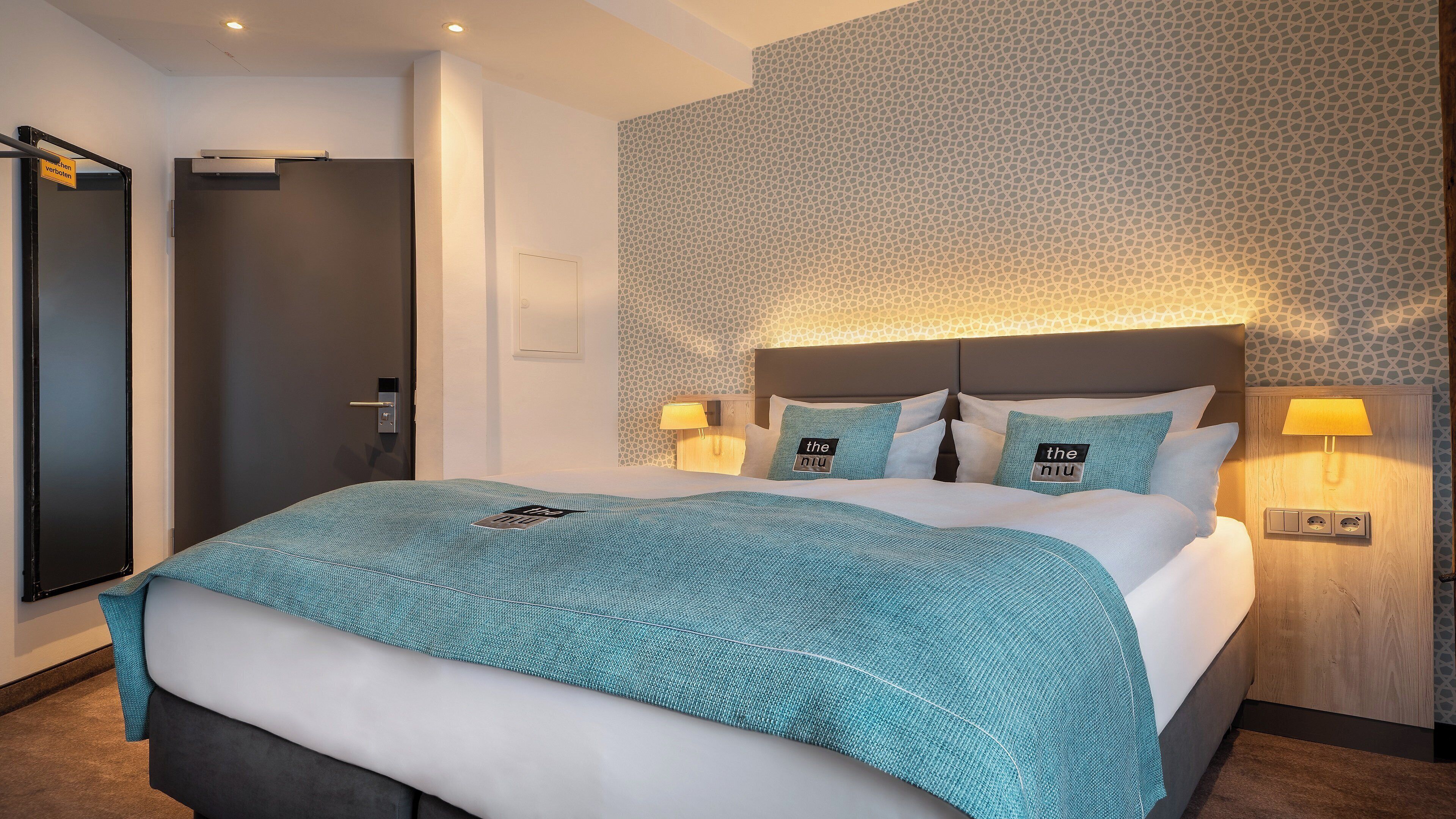 Une chambre d’hôtel avec un grand lit, deux lampes de chevet, une tête de lit en bois et un papier peint à motifs.
