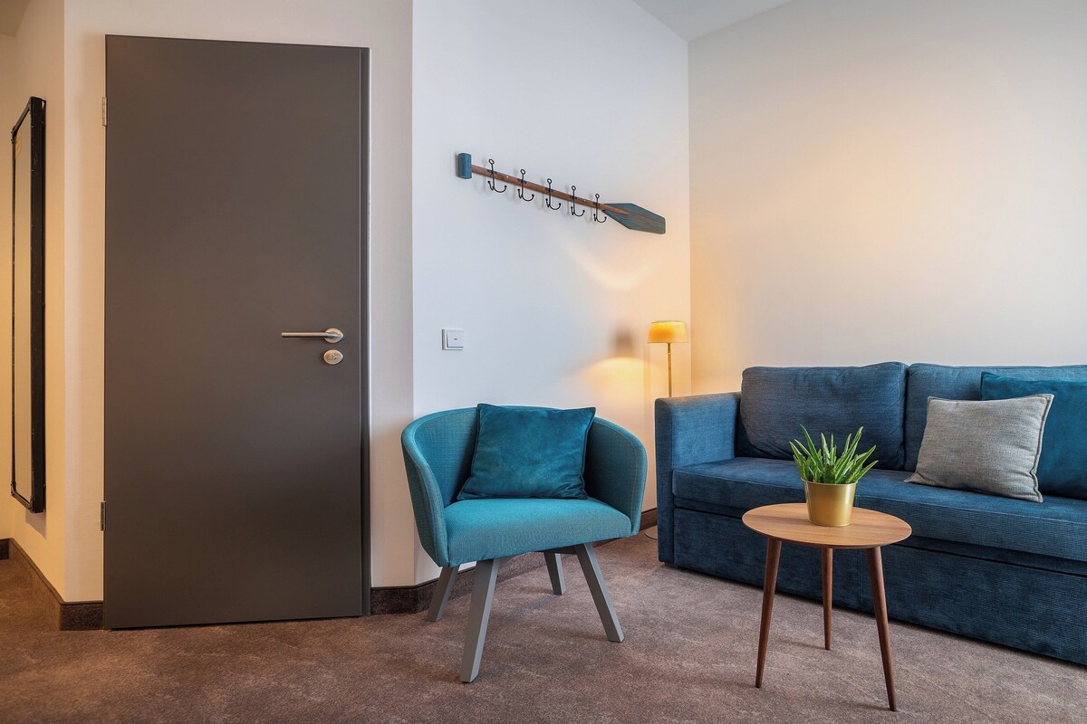 Una sala de estar moderna con una puerta oscura, un sofá azul con cojines, un sillón azul pequeño y una mesa redonda de madera con una planta.
