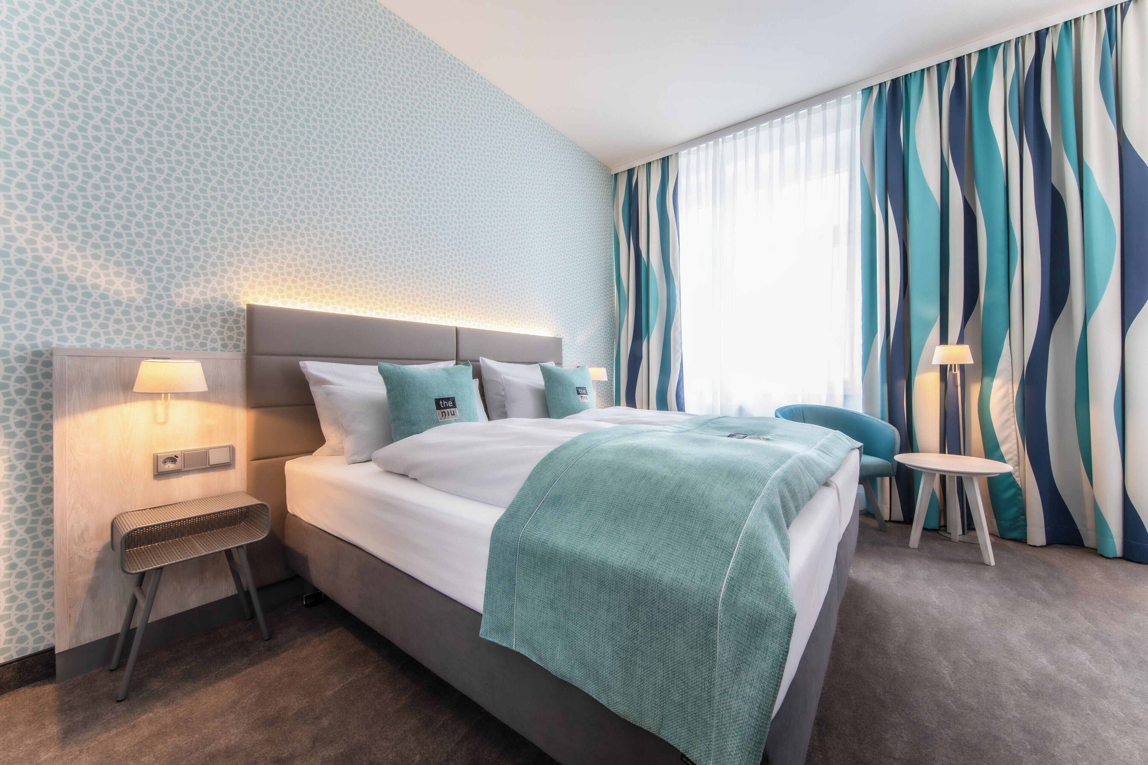 Una habitación de hotel moderna con una cama grande, una mesita de noche con lámpara, una pequeña mesa redonda y una silla.