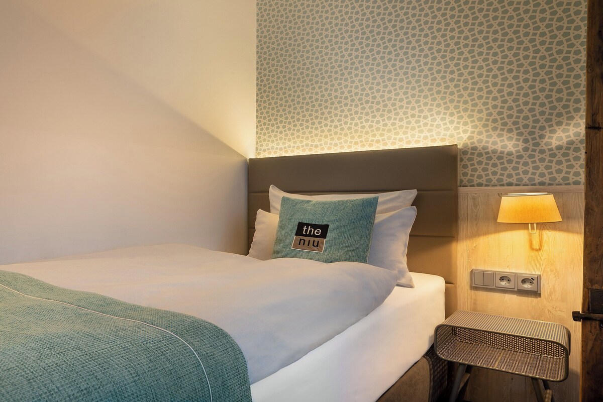 A neatly made bed with a green blanket, a pillow with text, a bedside lamp, and a small wooden bench.