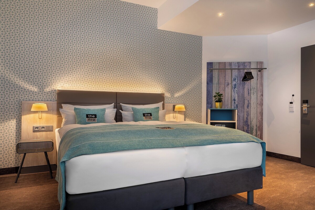 A modern bedroom with a large bed, two bedside tables with lamps, a small side table, and a wooden panel wall.