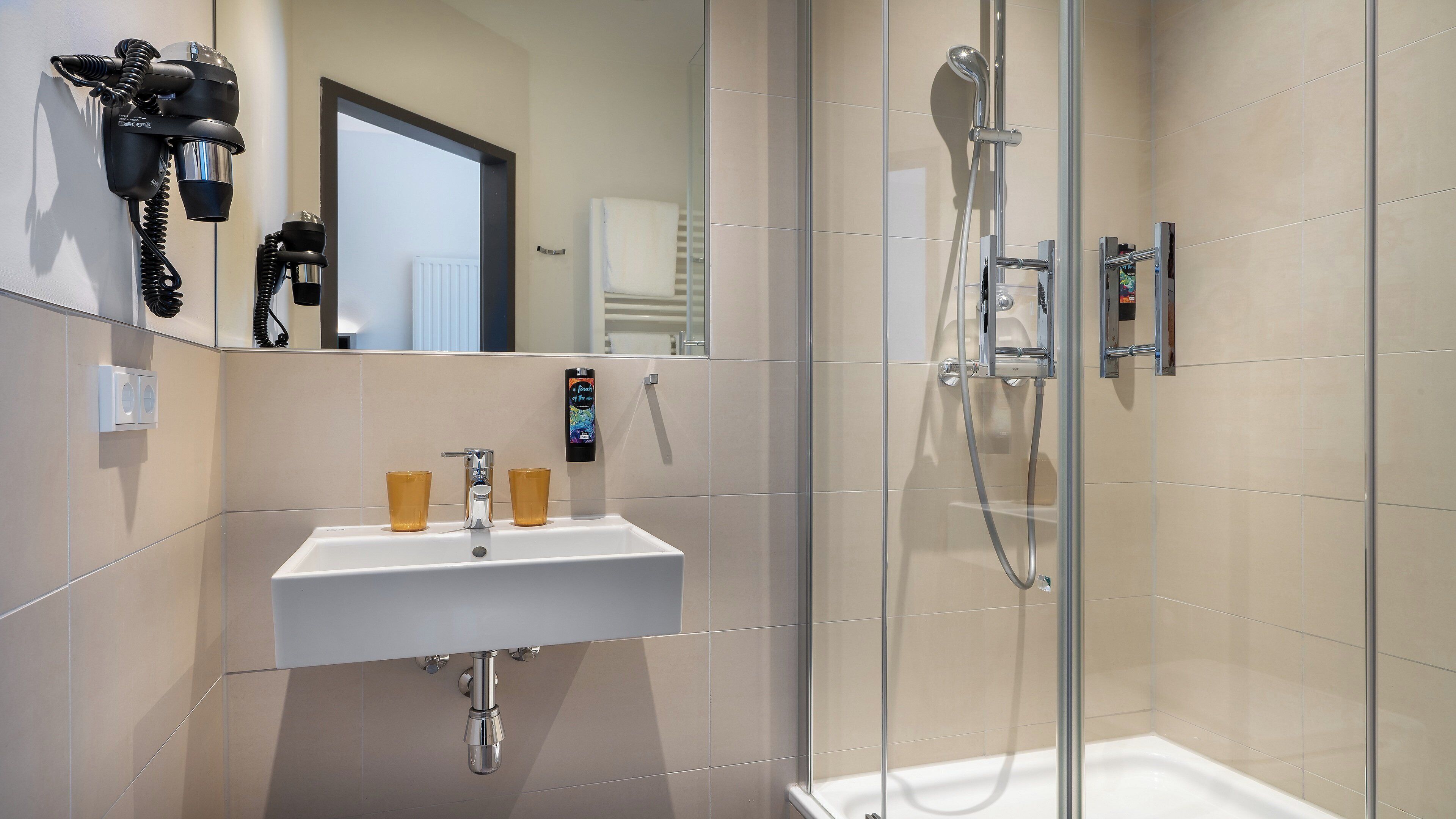 A modern bathroom with a glass-enclosed shower, a white sink with two cups, and a hairdryer mounted on the wall.