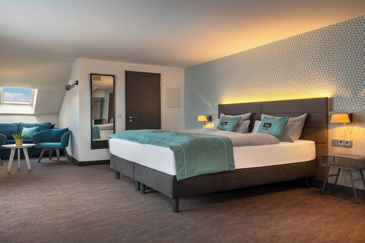A modern hotel room with a large bed, a sofa, a small table, and a mirror.