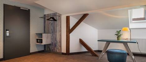 Una habitación moderna con una puerta oscura, estructura de vigas de madera, un pequeño escritorio con lámpara y una silla azul.