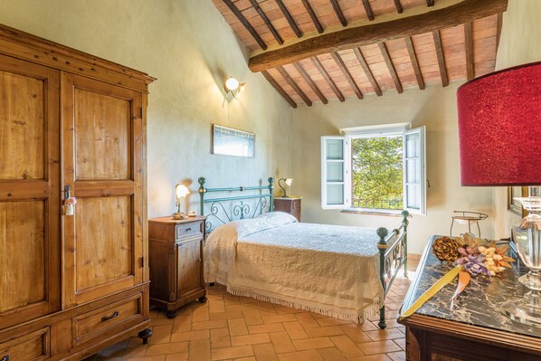 A bedroom with a wooden wardrobe, a bed with a white bedspread, a nightstand with a lamp, a window with a view of greenery, and a red chair. - Podere Albereto (Buonconvento)