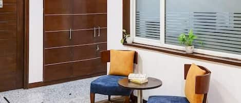 A room with a chandelier, a large window with blinds, a door, and two chairs with a small table.