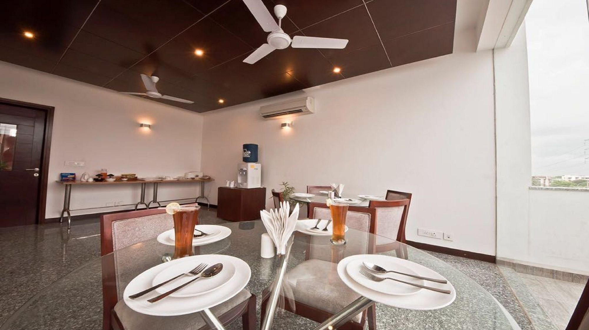 A modern dining area with a glass table, white plates, and cutlery. There are ceiling fans, recessed lighting, and an air conditioning unit.