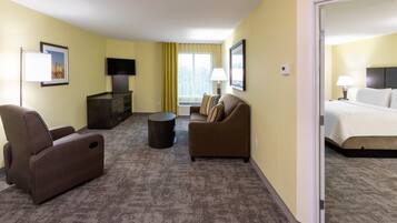 A hotel room with a sofa, armchair, and a bed. There is a television on the wall and a window with curtains.