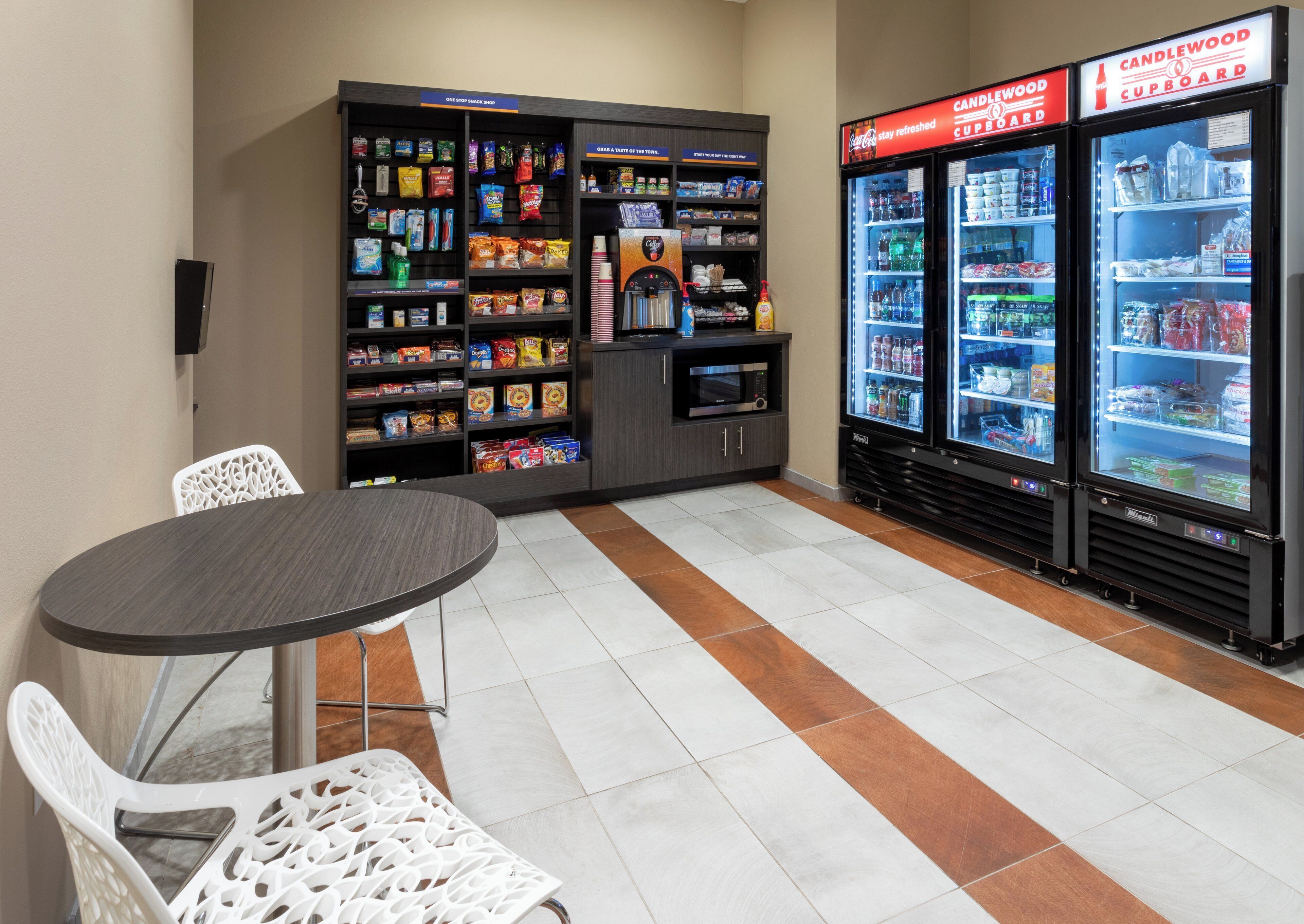 A snack shop with shelves stocked with various snacks and a microwave. There are two refrigerator freezers with beverages and snacks. A table with chairs is in the foreground.