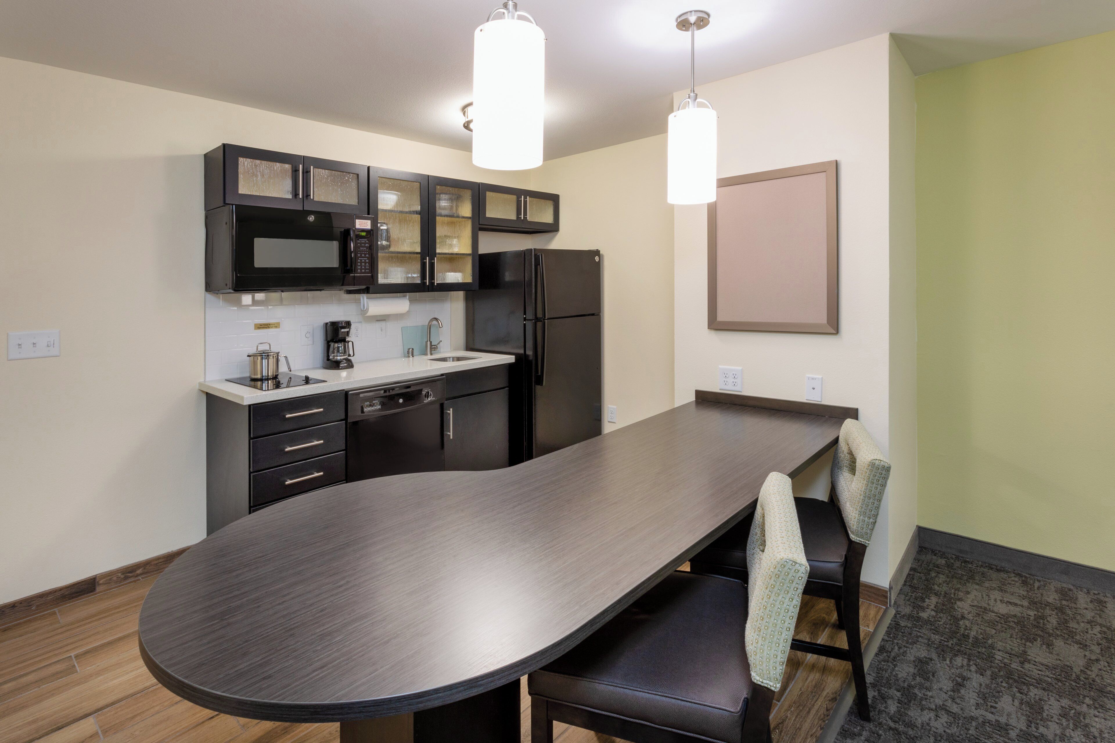 A modern kitchen with a dark countertop, black cabinets, a microwave, a refrigerator, and a dining area with a long table and chairs.