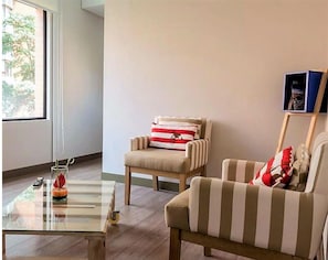 A room with a glass coffee table, two striped armchairs, a small table with a remote control and a decorative item, and a window with a view of trees.