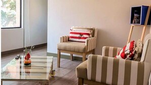 A room with a glass coffee table, two striped armchairs, a small table with a remote control and a decorative item, and a window with a view of trees. - Mika Suites (Bogotá)
