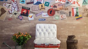 A room with a white cushioned chair, a potted plant, and a vintage suitcase. - Mika Suites (Bogotá)