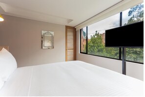 A hotel room with a large bed, a TV mounted on the wall, a window with a view of buildings and trees, and a decorative mirror on the wall. - Mika Suites (Bogotá)