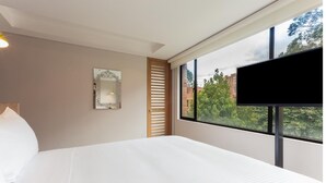 A hotel room with a large bed, a TV mounted on the wall, a window with a view of buildings and trees, and a decorative mirror on the wall. - Mika Suites (Bogotá)