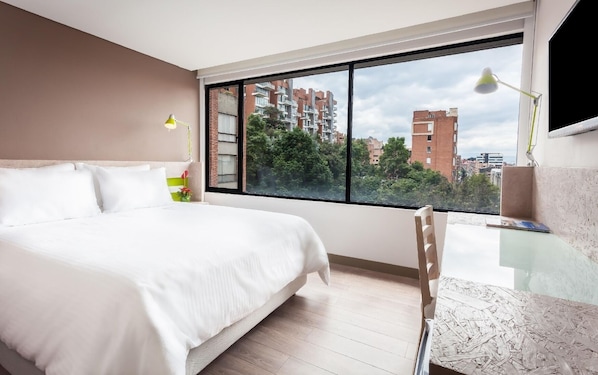 A modern hotel room with a large bed, a desk, and a view of buildings and trees. - Mika Suites (Bogotá)