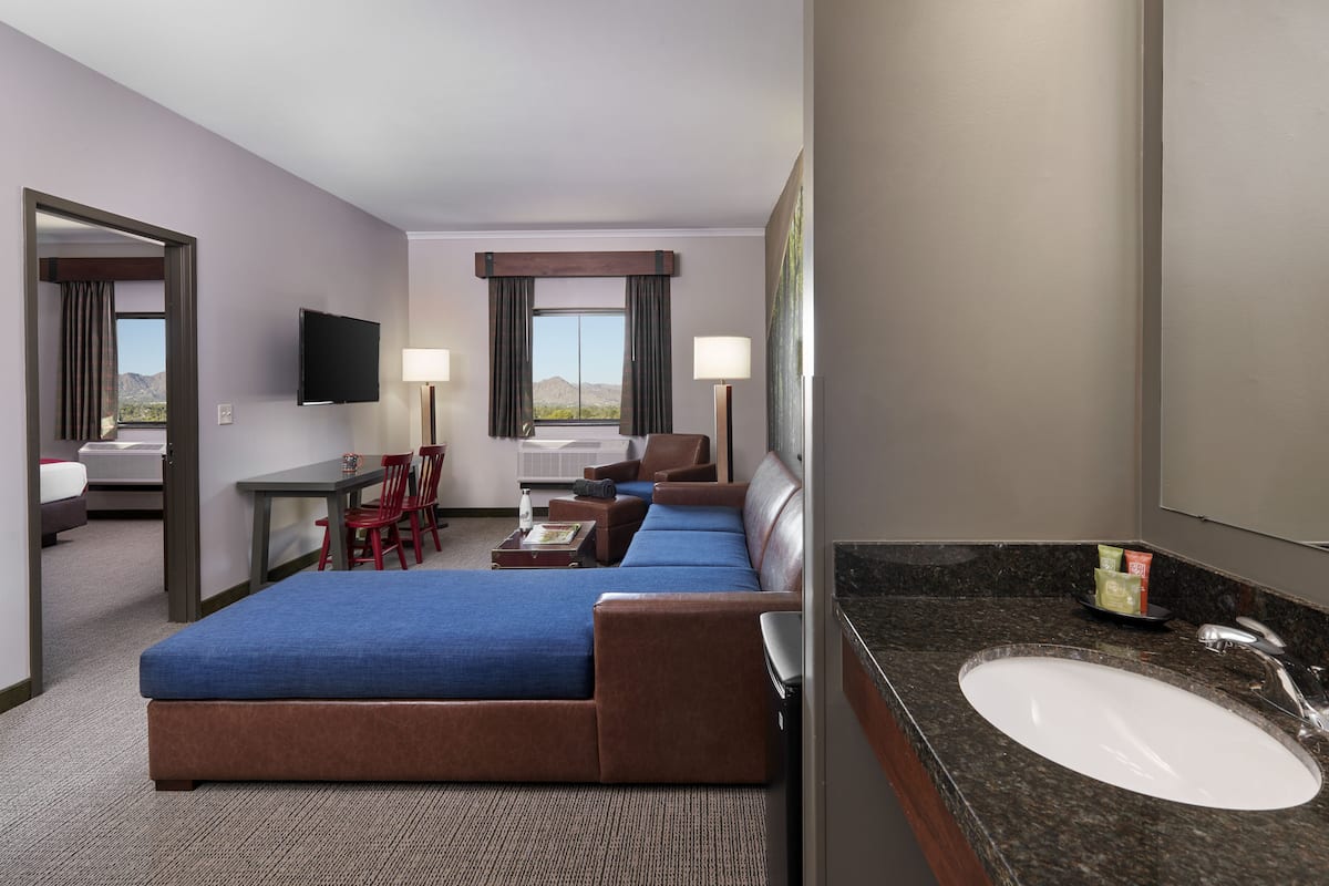 Habitación de hotel con cama, escritorio, dos sillas, sofá, televisión, lámpara y baño con lavabo y espejo.