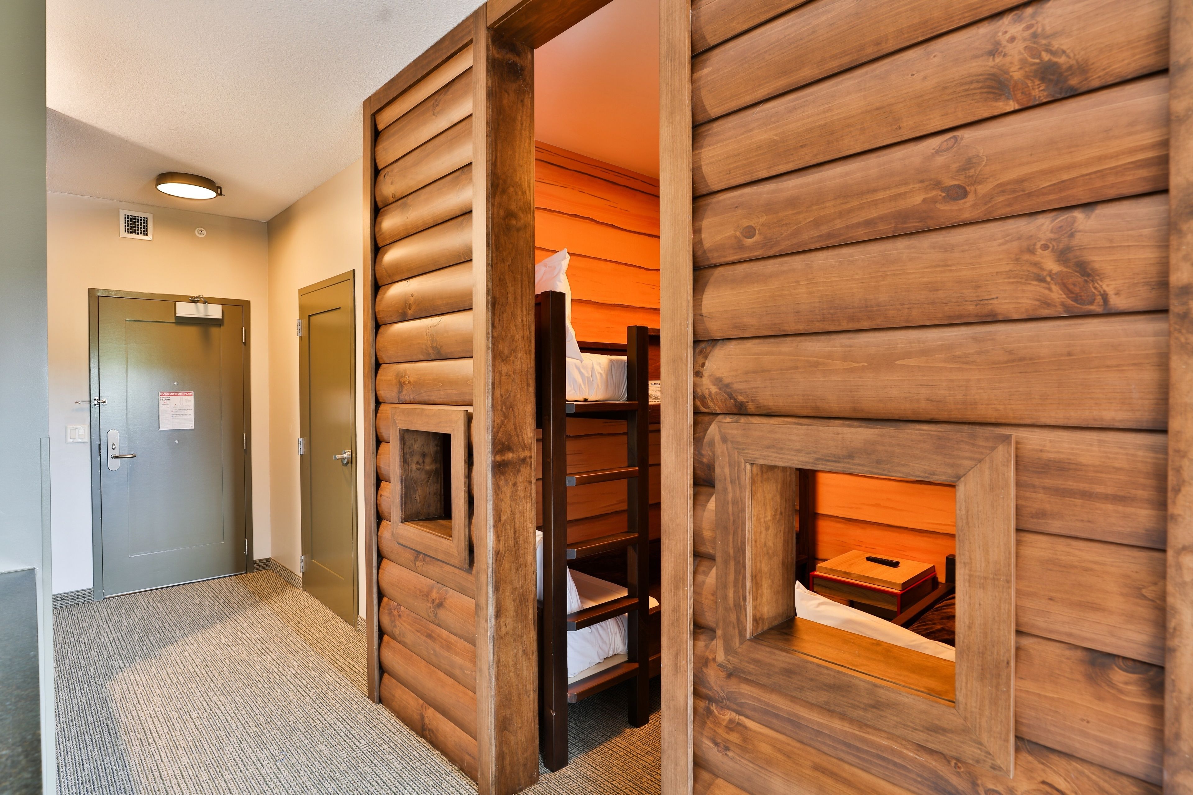 A hotel room with a wooden log wall, a bed with a headboard, a small wooden chest, and a door with a sign.