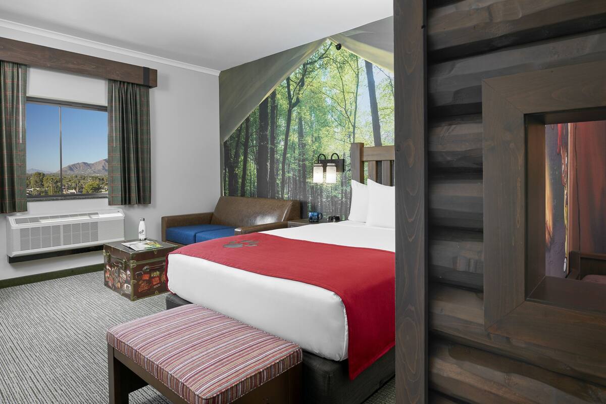 Habitación de hotel con una cama grande, un escritorio, una silla, un ventanal con cortinas, un radiador y un cuadro de una escena de bosque.