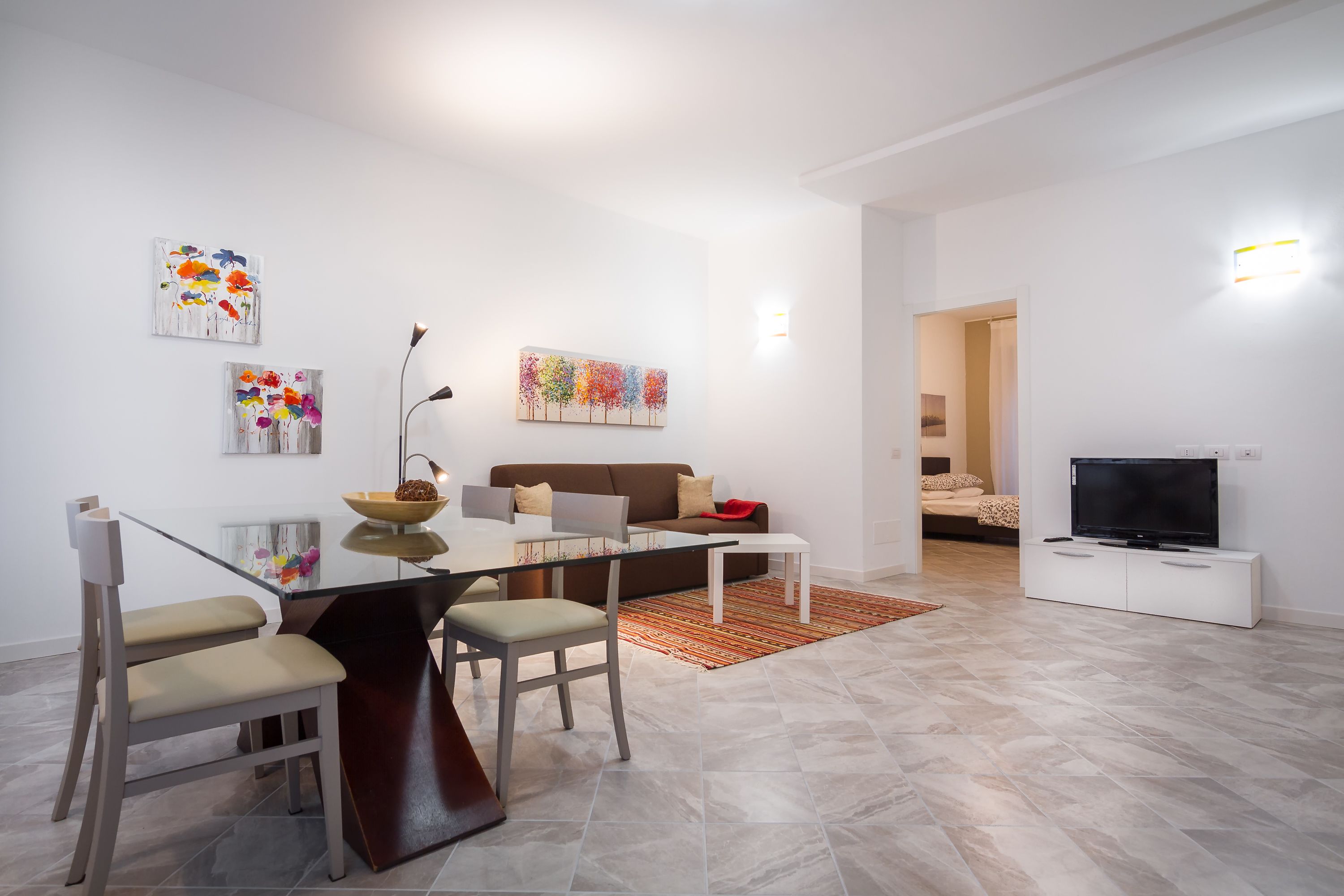 A modern living room with a glass dining table, a sofa, a TV, and artwork on the walls.