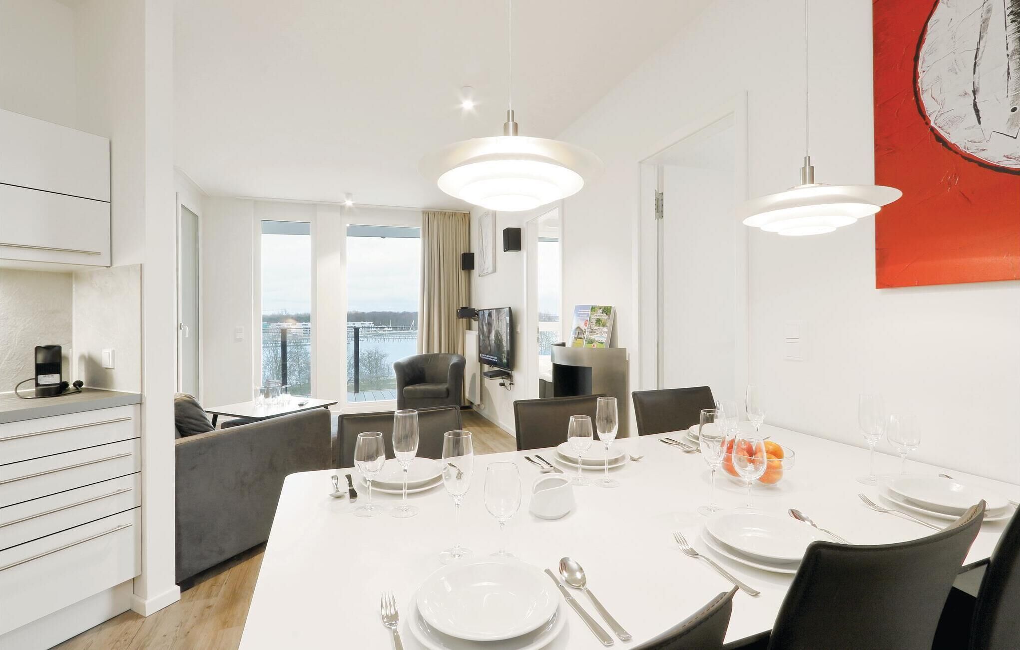 Un comedor moderno con una mesa blanca para cuatro, ventanas amplias con vista a un cuerpo de agua y un cuadro abstracto rojo en la pared.