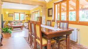 Un espace repas comprenant une table et des chaises en bois, un lustre et de grandes fenêtres à cadre en bois.