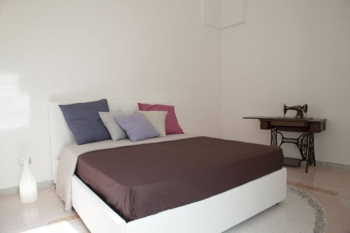 Une chambre à coucher avec un lit, des oreillers, un bureau avec une machine à coudre et un mur blanc.