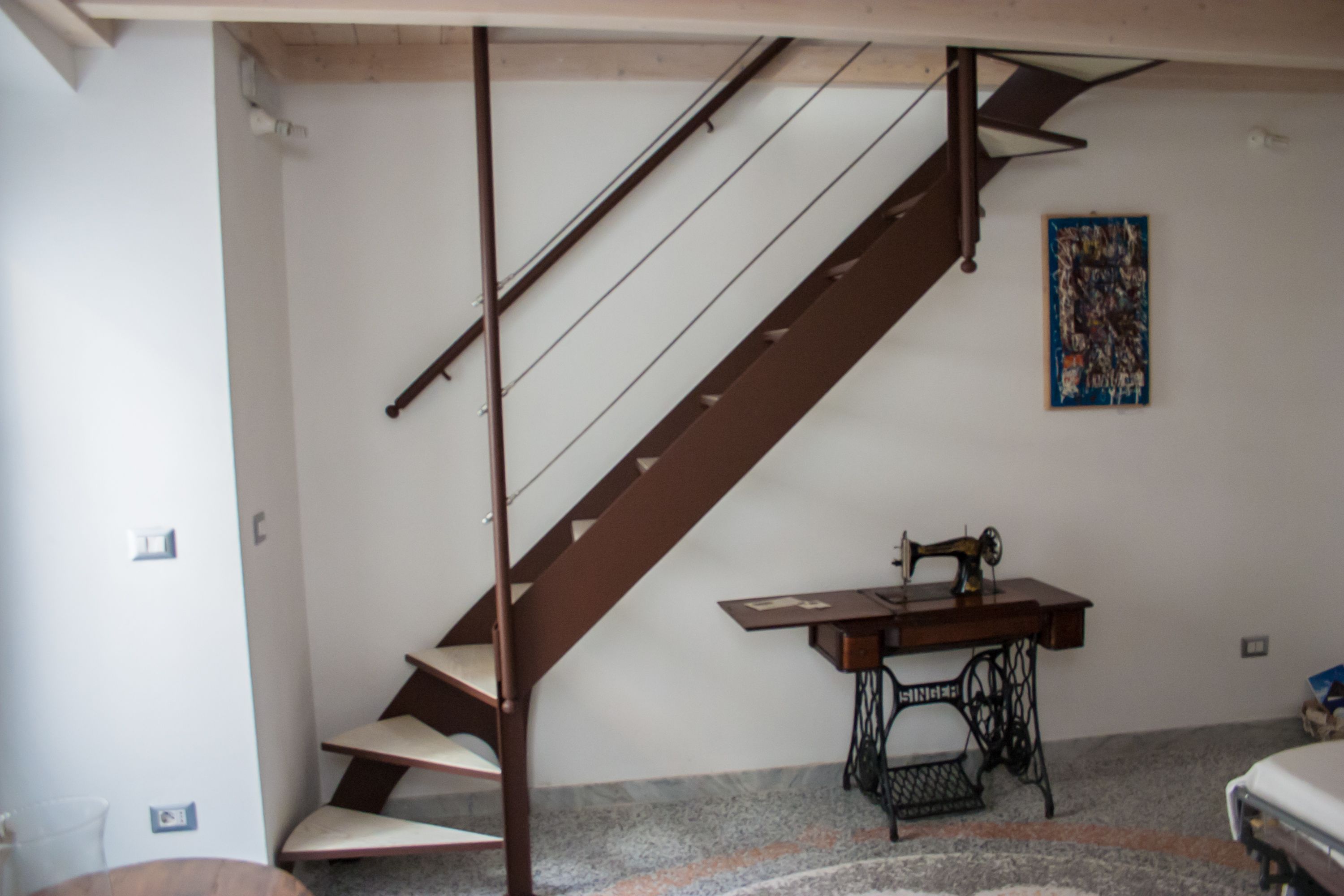Une pièce avec un escalier, une machine à coudre et un tableau accroché au mur.