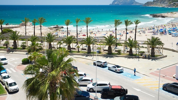 En parkeringsplads ved stranden med biler, palmer og udsigt over strand og hav.