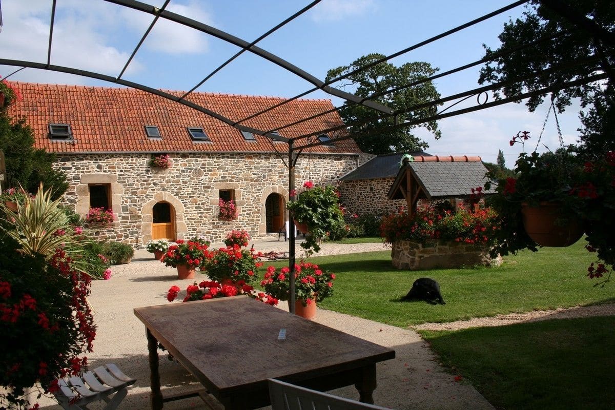 A stone building with a red-tiled roof, a covered patio area with a table and chairs, and a well-maintained garden with red flowers.