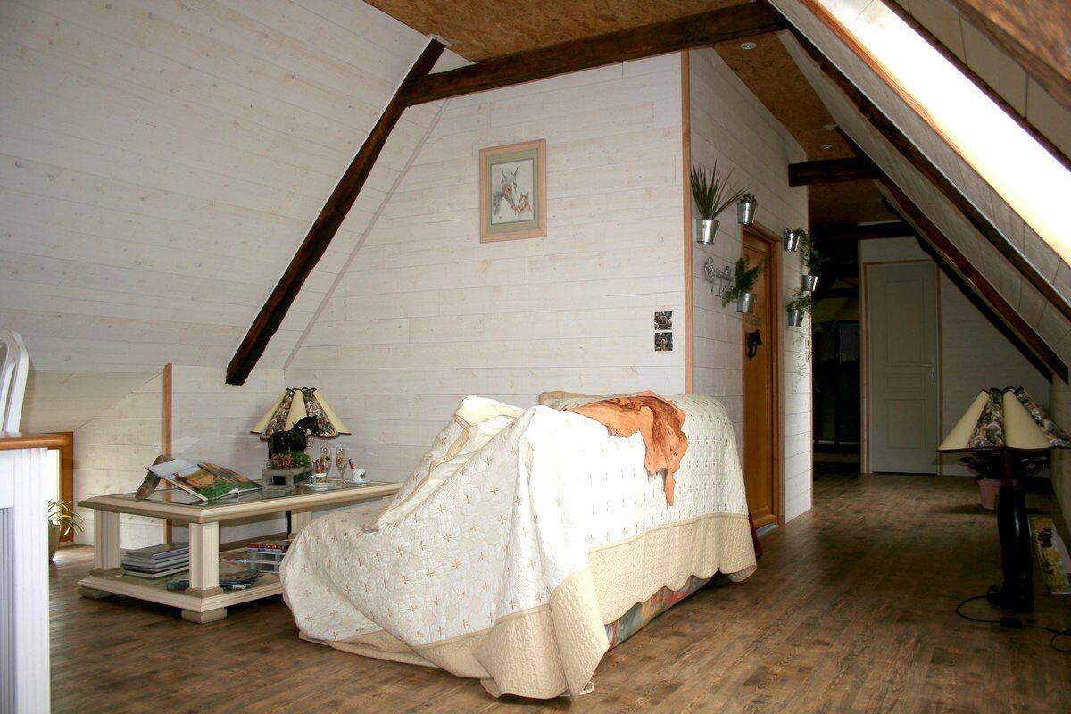 A cozy attic bedroom with a sloped ceiling, a bed with a white bedspread, a small table with books, and a lamp.