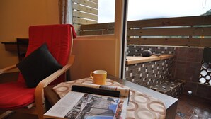 Una habitación con una silla roja, una mesa con un periódico, una taza amarilla y vistas a un patio empedrado.