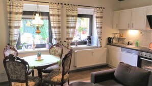 A dining area with a table and chairs, a kitchen with appliances, and a window with curtains. - Müller's Landidyll - Apartments Müller's Landidyll (Klitten)