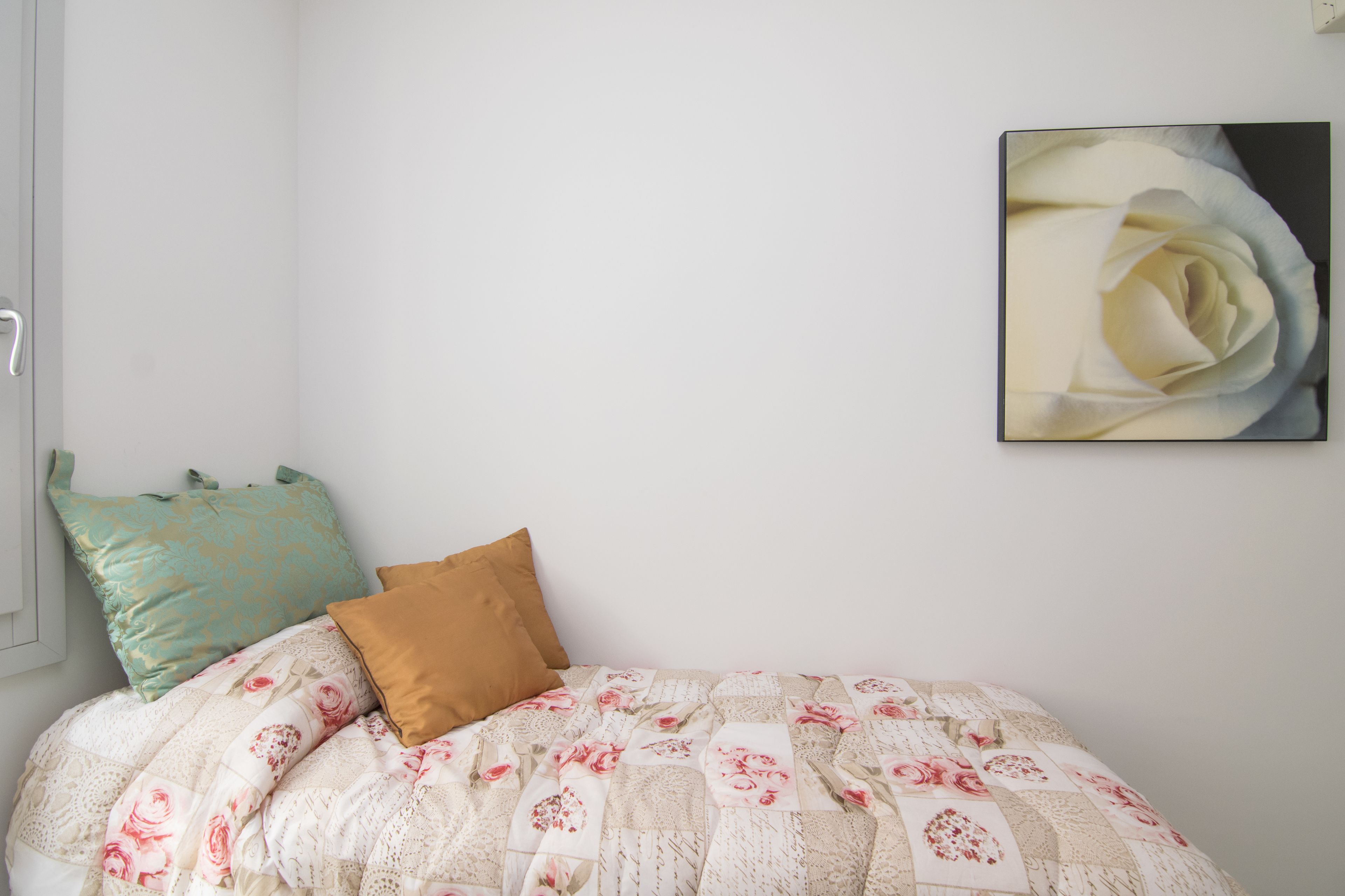 A bedroom with a bed, two pillows, a wall-mounted picture of a rose, and a window with a white frame.