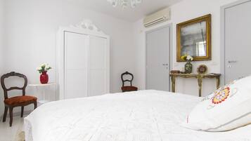 Un dormitorio con una cama grande, una mesita con un florero, dos sillas y un espejo en la pared.