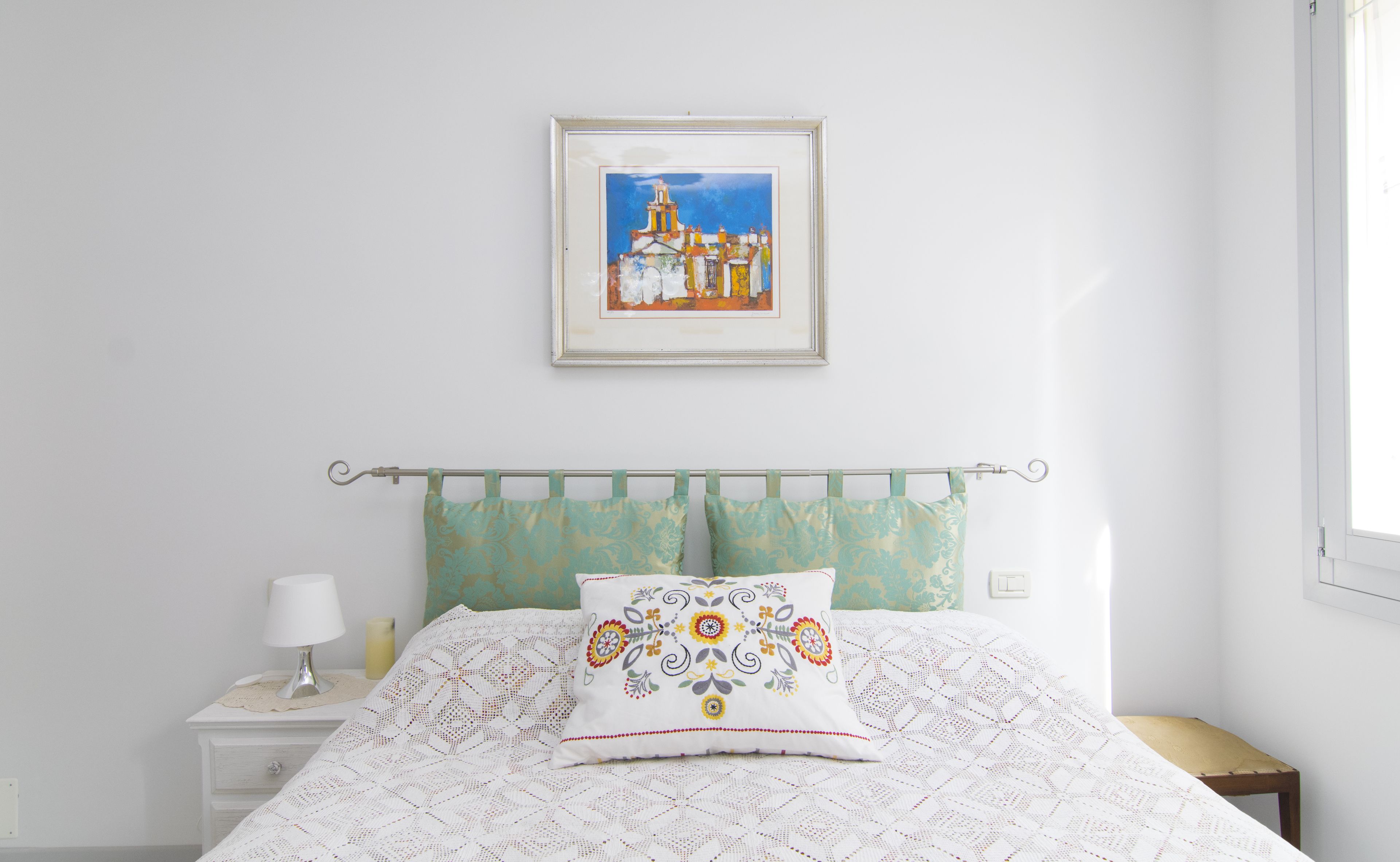 A neatly made bed with a patterned pillow, a bedside lamp, and a framed painting of a building on the wall.