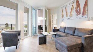 A spacious living room with a grey sofa, armchairs, and a coffee table. A large window offers a view of the city. - HighEnd Travemünde - Whg 201 (Lübeck Travemünde)