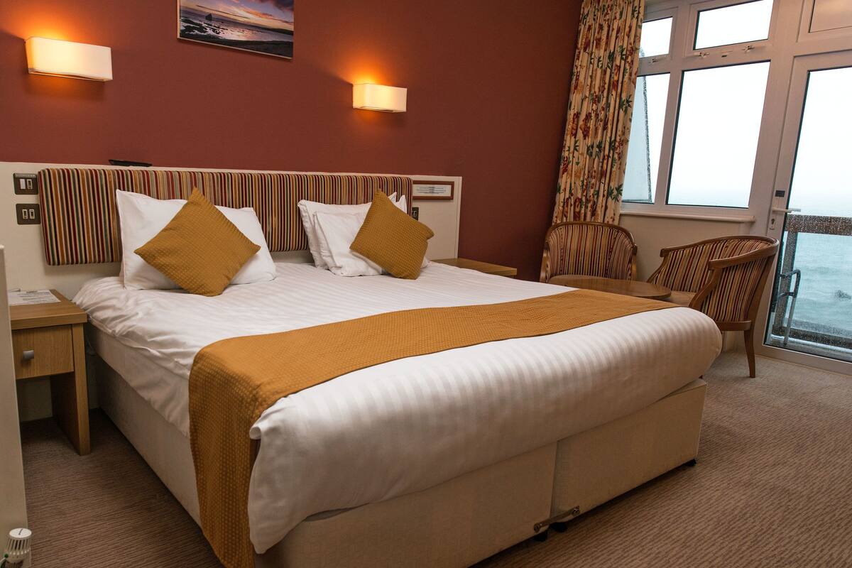 A hotel room with a large bed, a wooden nightstand, a chair, and a window with curtains.