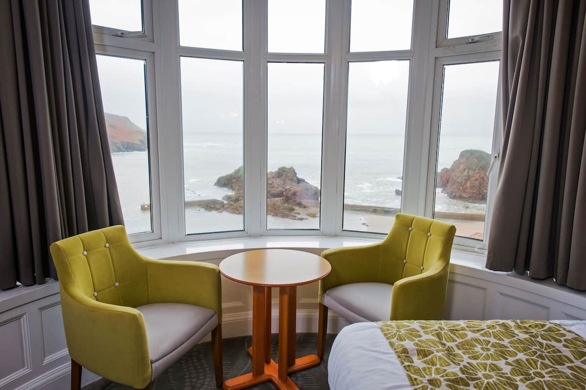 A hotel room with a large window offering a view of the sea and rocky outcrops. There are two yellow armchairs and a small round table.