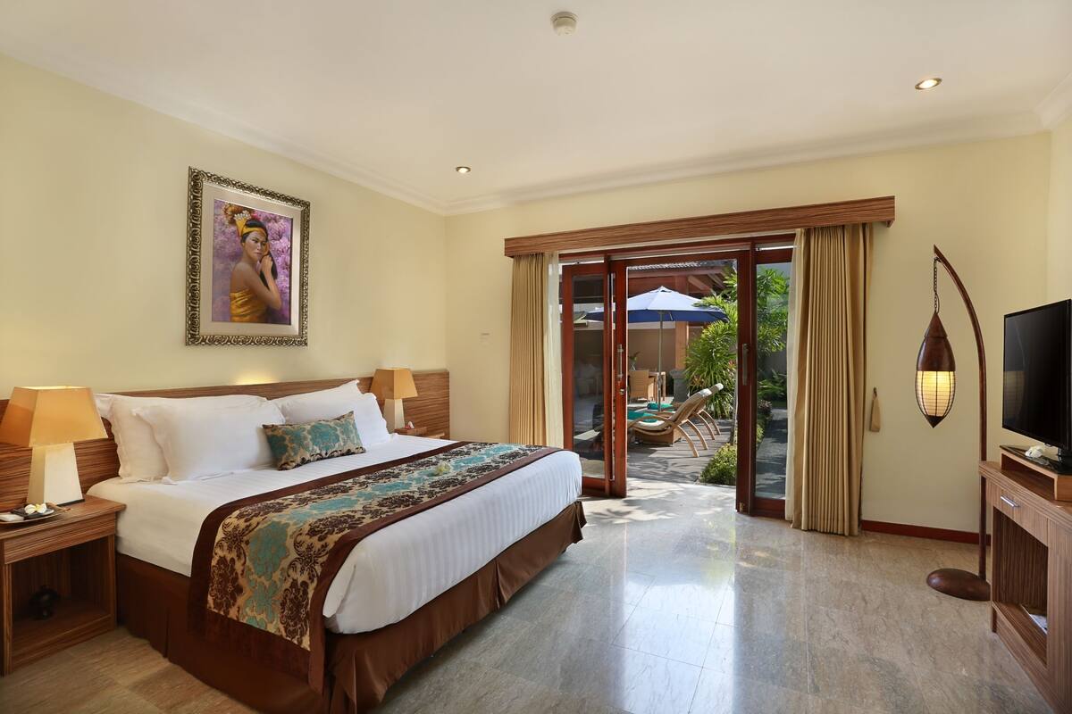 A hotel room with a large bed, a TV, a nightstand, and a view of the outdoor area.