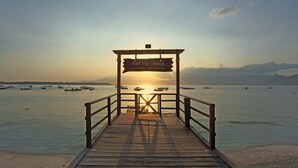 A wooden pier leading to a sign for Hotel Vila Ombak, with boats on the water and mountains in the background. - Akoya Pool Villa (Gili Trawangan)
