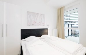 A hotel room with a bed, a headboard, a window with curtains, a wall-mounted artwork, and a door. - HighEnd Travemünde - Whg 05 (Lübeck Travemünde)