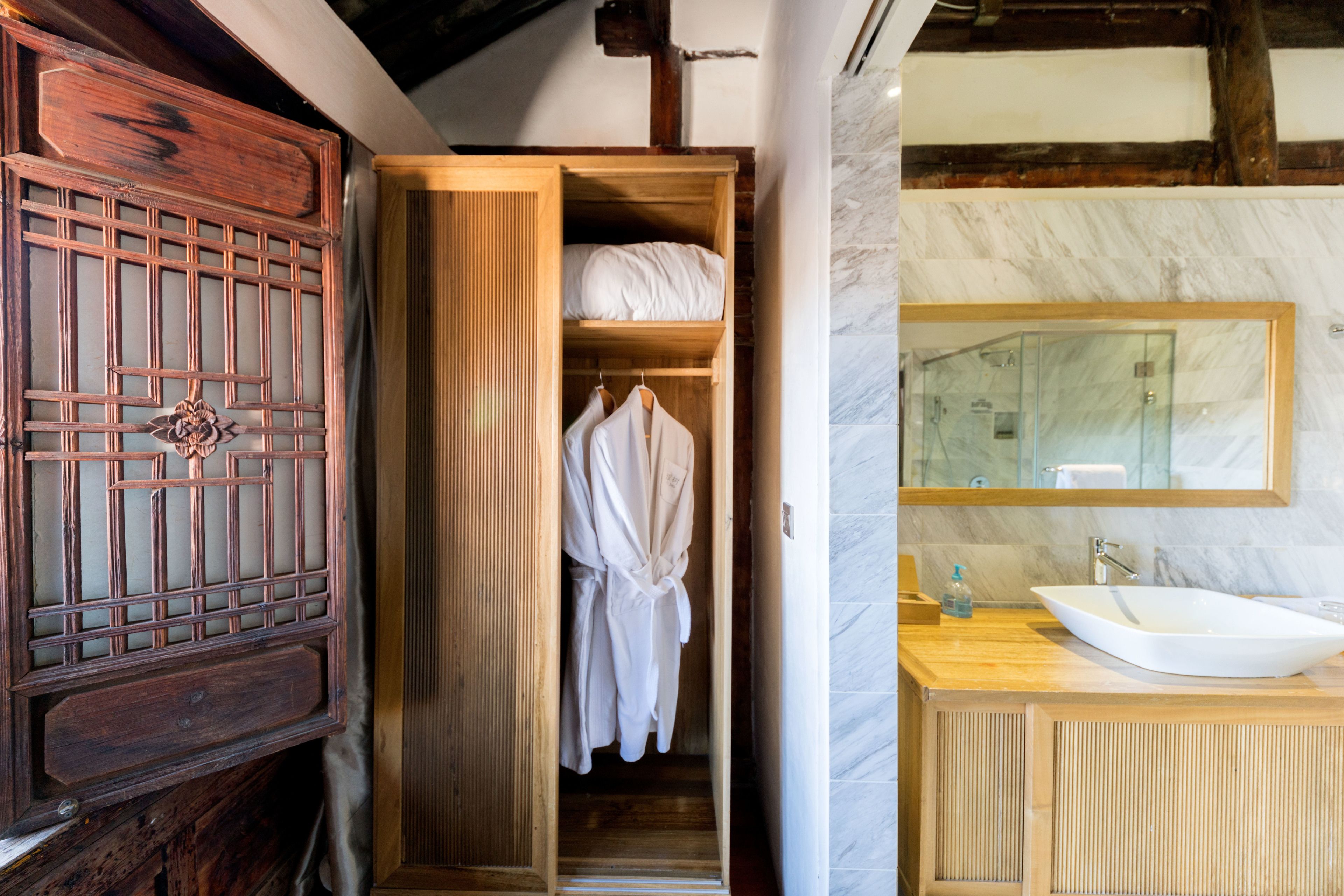 A wooden wardrobe with a traditional design, a robe hanging inside, and a modern bathroom with a marble sink and a large mirror.