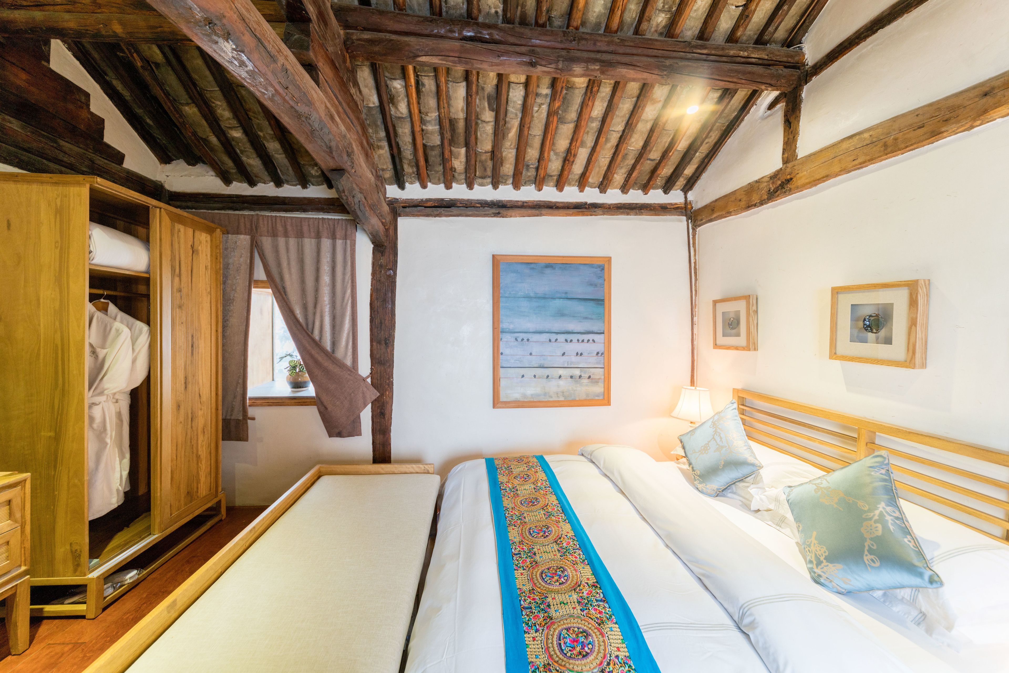 Un dormitorio con techo de madera, una cama con colcha bordada de colores, un armario de madera y una ventana con cortinas.