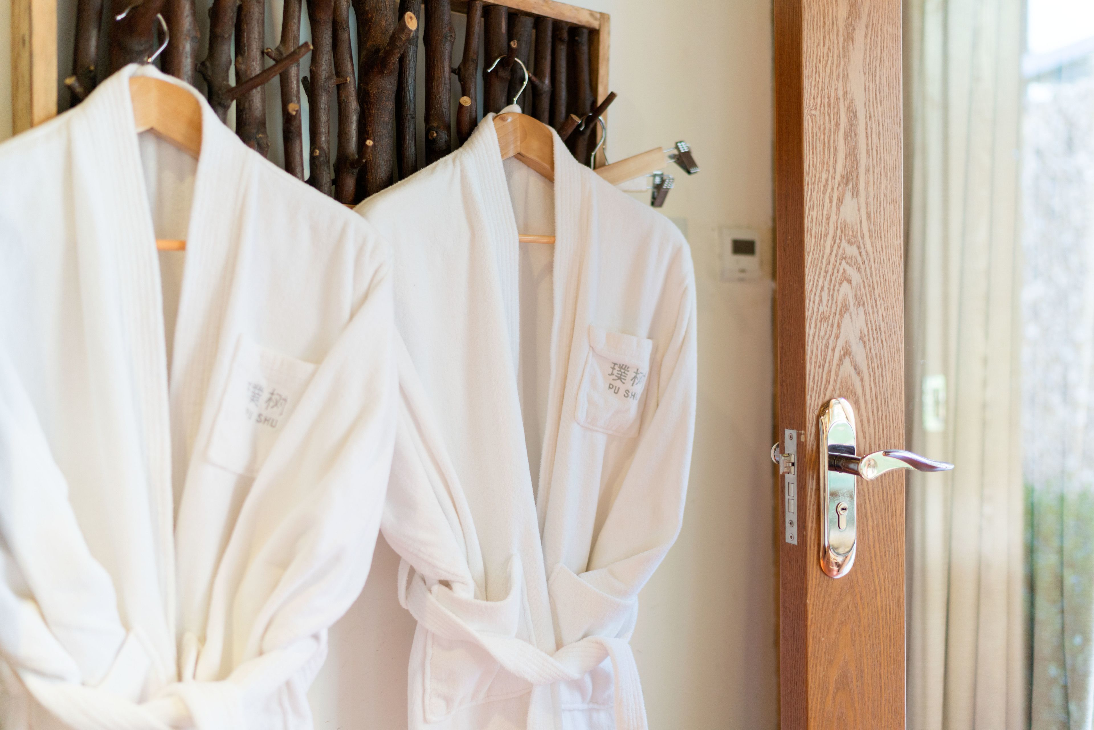 Deux robes blanches sont suspendues à des cintres en bois dans une pièce dotée d’une porte en bois et d’une fenêtre avec des stores.