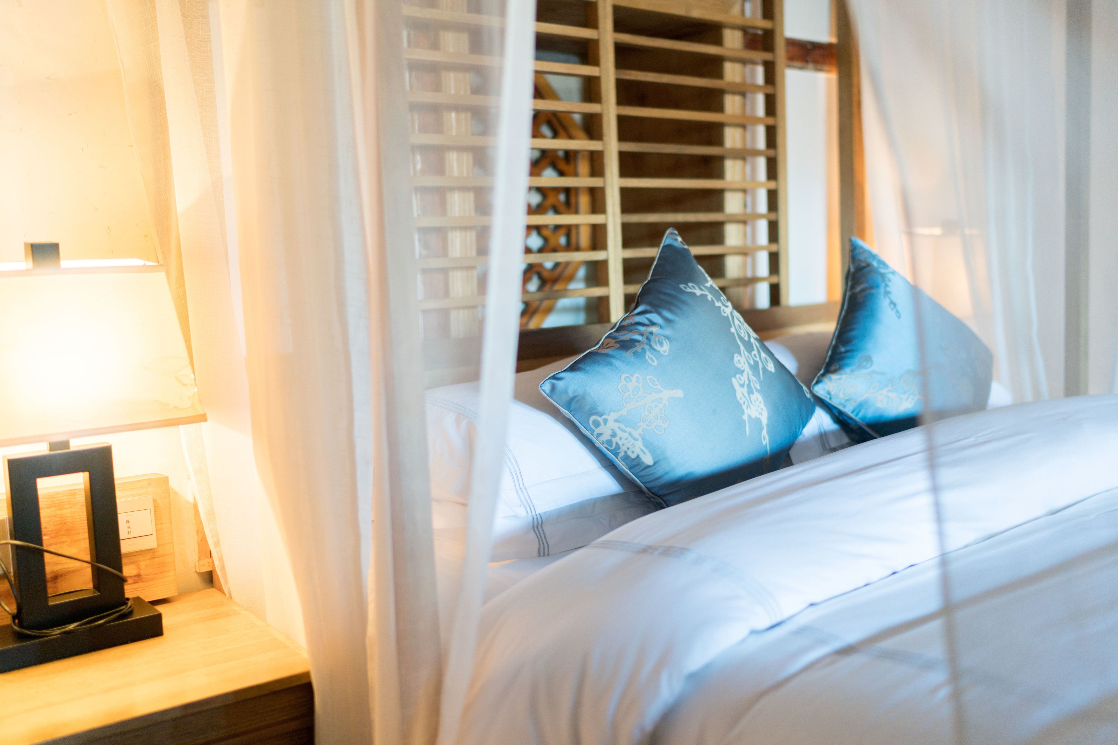 A neatly made bed with white linens and two blue pillows, a bedside table with a lamp, and sheer curtains.