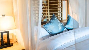 A neatly made bed with white linens and two blue pillows, a bedside table with a lamp, and sheer curtains.