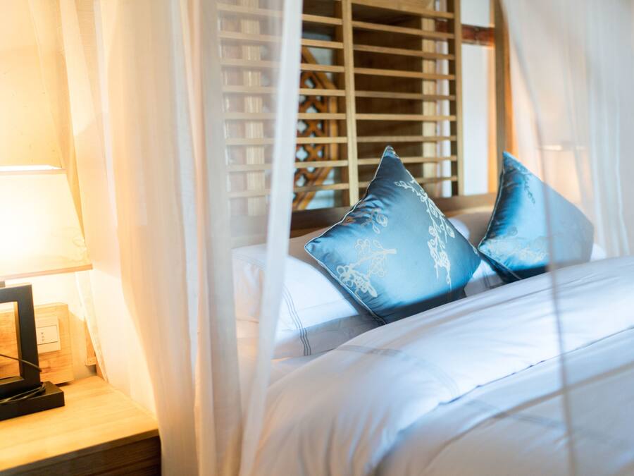 A neatly made bed with white linens and two blue pillows, a bedside table with a lamp, and sheer curtains.