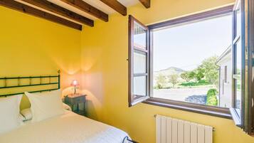A bedroom with a bed, a nightstand with a lamp, a window with a view of the outside, and wooden beams on the ceiling.